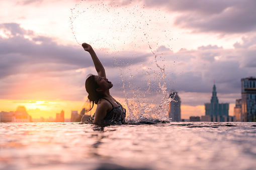 Black silhouette of asian woman splash water on summer vacation holiday relaxing in infinity swimming pool with blue sea sunset view and building urban city background. Healthy happiness lifestyle