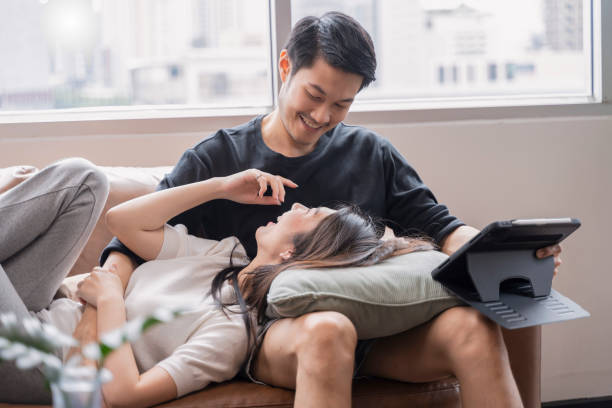 lifestyle at home,asian female stay home together with her husband watch movie online by tablet on sofa in living room at home,sweet asian marry couple spending weekend time together at home stock photo