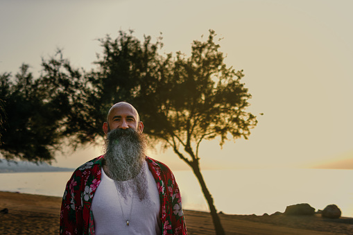 Headshot portrait of a hawaiian bearded man with shaved head standing on a beach with a tree while the sun comes up through the sea horizon