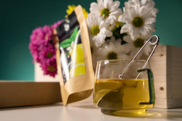 Glass cup with tea and tea strainer, flowers, herbs and wooden\ncrate on the white table behind teal background