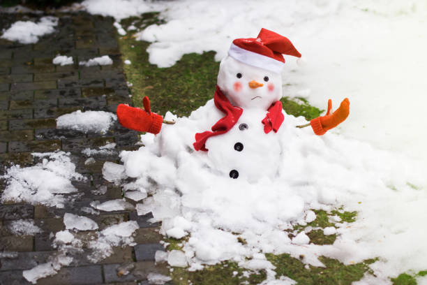 Unhappy snowman in mittens, red scarf and cap is melting  outdoors in sunlight stock photo