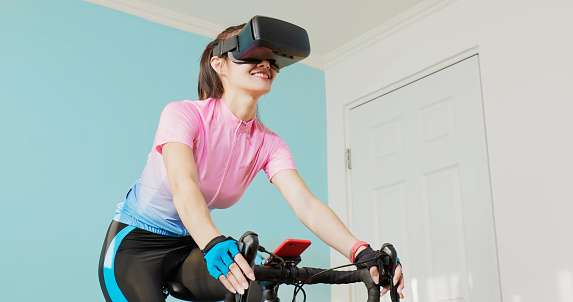 woman cycling with VR glasses in her room - asian female cyclist is playing online bike games