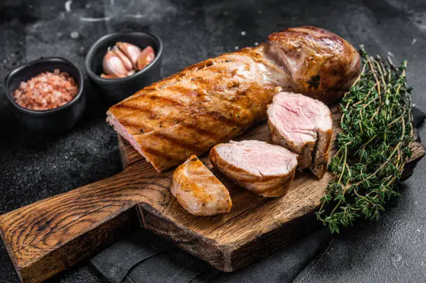 BBQ roasted pork tenderloin fillet meat on wooden board with herbs. Black background. Top view.