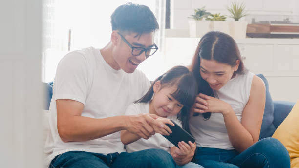 счастливая азиатская семья папы, мамы и дочери играет в забавную игру онлайн на смартфоне сидит диван в комнате дома. самоизоляция, пребыва� - ipad iphone smart phone ipad 3 стоковые фото и изображения