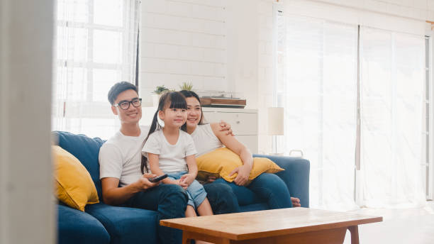 happy asian family enjoy their free time relax together at home. lifestyle korean dad, mom and daughter watching tv together and having fun lying on sofa in living room in modern house. - apartment television family couple imagens e fotografias de stock