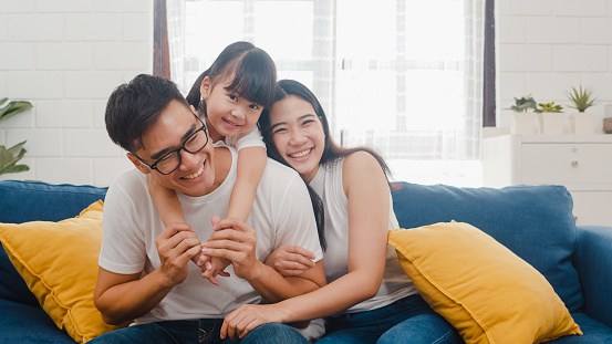 Happy cheerful Asian family dad, mom and daughter having fun cuddling and video call on laptop on sofa at house. Self-isolation, stay at home, social distancing, quarantine for coronavirus prevention.