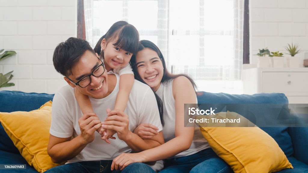 Happy cheerful Asian family dad, mom and daughter having fun cuddling and video call on laptop on sofa at house. Self-isolation, stay at home, social distancing, quarantine for coronavirus prevention. Happy cheerful Asian family dad, mom and daughter having fun cuddling and video call on laptop on sofa at house. Family Stock Photo