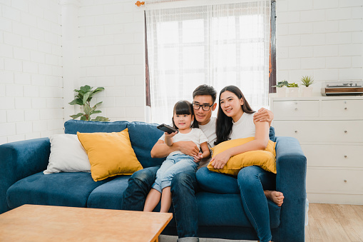 Lifestyle Korean dad, mom and daughter watching TV together and having fun lying on sofa in living room in modern house.