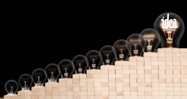 Group of light bulbs going from dark one to a shining one with fiber in Idea shape on wooden block ladder isolated on black background. Concept of Thinking, Success and Creativity.