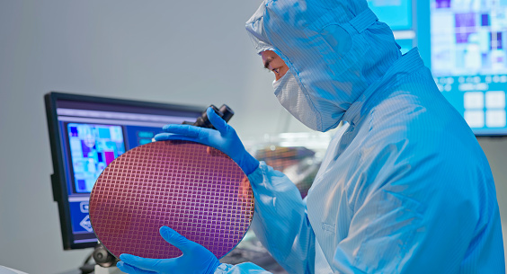 asian male technician in sterile coverall holds wafer with gloves and check it at semiconductor manufacturing plant