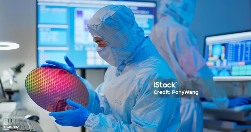 technician with wafer asian male technician in sterile coverall holds wafer that reflects many different colors with gloves and check it at semiconductor manufacturing plant Semiconductor Stock Photo
