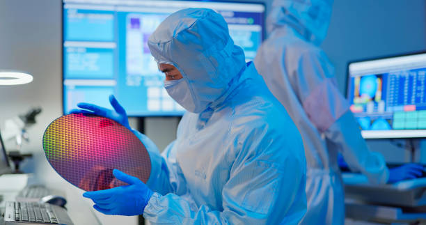 technician with wafer asian male technician in sterile coverall holds wafer that reflects many different colors with gloves and check it at semiconductor manufacturing plant cleanroom stock pictures, royalty-free photos & images