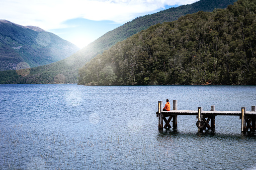 Young man sitting on the pier, surrounded by a beautiful landscape. Photo taken from a distance