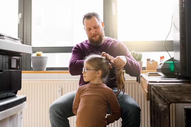 Man working at home and making a ponytail to his daughter Mature man making a ponytail to his daughter while working from home during pandemic. Man sitting at home office and taking care of his daughter. brushing hair stock pictures, royalty-free photos & images