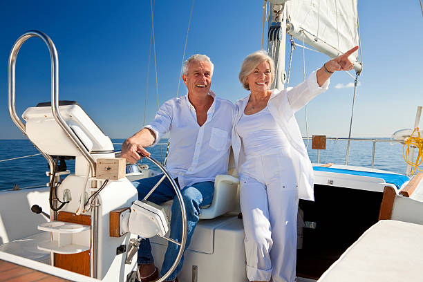 Happy Senior Couple At The Wheel of a Sail Boat A happy senior couple sitting at the wheel of a sail boat on a calm blue sea sailing couple stock pictures, royalty-free photos & images