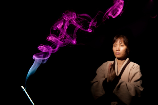 Asian woman in traditional clothes meditates in the background while facing colorful incense smoke.