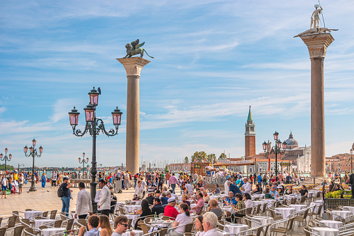 View over cafe at the famous San Marco square in Venice, Italy, with two symbols of Venice, winged lion and Saint Theodore killing a dragon, at sunset with many tourists