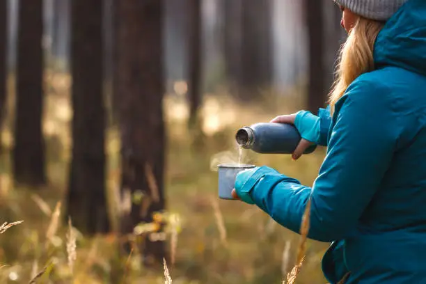 Hiker pouring hot drink from thermos into travel mug during trekking in woodland. Adventure in nature