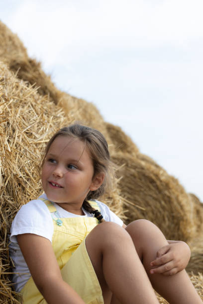 istock Portrait of curious little girl sitting on haystack with another stack behind back looking away with interested eyes with dozens of haystacks in background. Away from city 1363632148