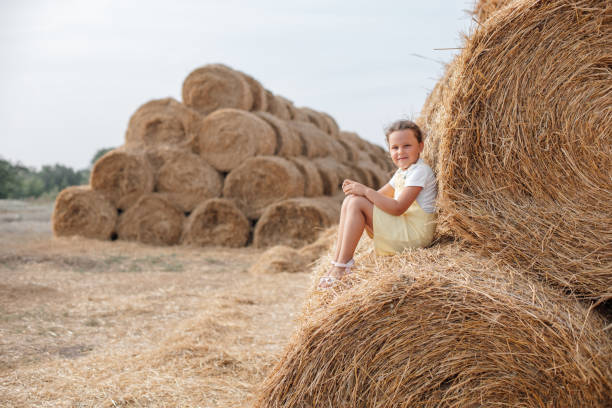 istock Happy female kid in sundress seating calmly on rolled haystack leaning on another stack with heap of haystacks in background resembling roof. Having fun away from city on field full of golden hay 1363637277