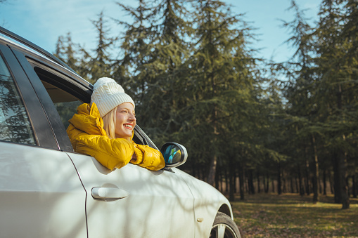 Happy young woman enjoying road trip through countryside, taking a break, leaning out of car window and smiling to perfect view in beautiful nature. Woman on the road trip