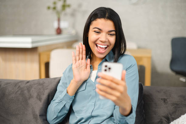 istock Cheerful young multiracial woman using cell phone for video call while sitting down on the comfortable couch at home. Smiling female student waving with hand while chatting with classmates 1363654074