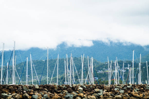 Masts of yachts berthed in the marina protected by a rock wall and with a low cloud mountain background scattered with homes, Airlie Beach, Queensland, Australia