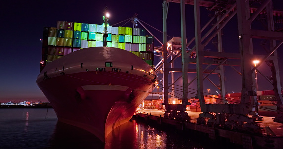 Low drone shot of a massive and heavily laden cargo ship docked at the Port of Long Beach at night, taken at the end of 2021 during the global supply chain crisis. The yard is visibly at capacity and there are multiple ships waiting for a berth out at sea.