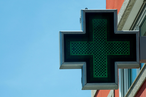 Pharmacy symbol green cross seen from the street sidewalk, apartment building facade, clear blue sky background. Copy space on the left. Galicia, Spain.