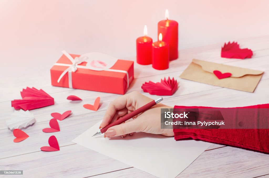a woman's hand writes a love letter for Valentine's Day. An envelope with a heart, a gift and candles. a woman's hand writes a love letter for Valentine's Day. An envelope with a heart, a gift and candles. High quality photo Writing - Activity Stock Photo