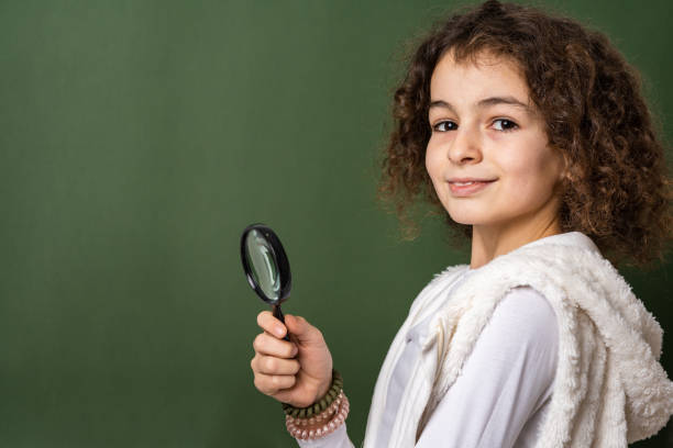 one small caucasian girl ten years old with curly hair front view portrait close up standing in front of green background looking to the camera holding magnifying lens education and learning concept - magnifying glass lens holding europe imagens e fotografias de stock