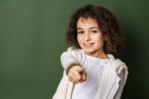 one small caucasian girl ten years old with curly hair front view portrait close up standing in front of green background looking to the camera pointing finger copy space smiling happy and joy - 10 11 years child human face female imagens e fotografias de stock