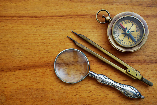 Directly above shot of a vintage brass compass, compasses and magnifying glass on wooden background.