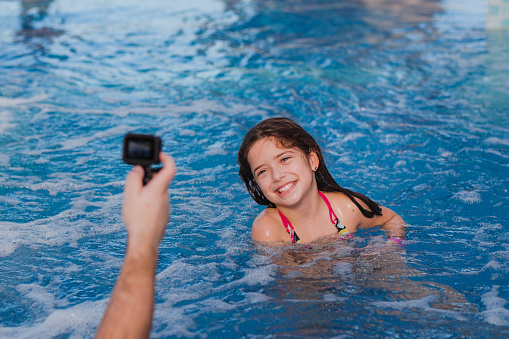 Cheerful young girl with a toothy smile swimming when parent is using go pro in order to take a photo of her