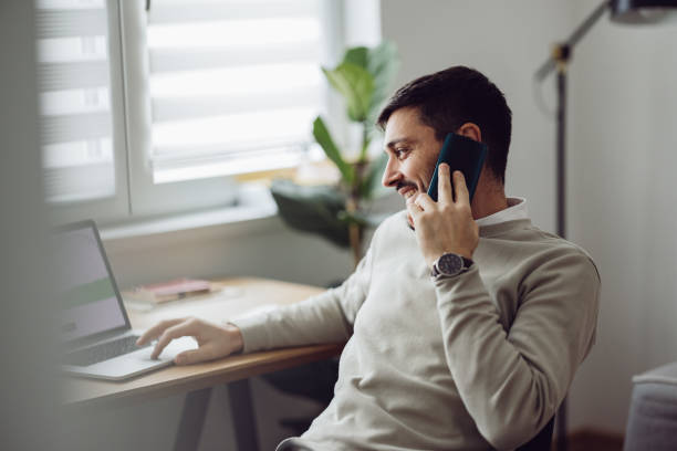 Home Office: An Anonymous Man Using a Laptop for Online Trading While Working From Home Caucasian businessman is talking on his smartphone while using a laptop for online trading and following the stock market from home. It's morning or later in the day. happy using phone stock pictures, royalty-free photos & images