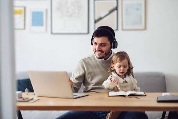 Home Office: Man Having a Video Call and Holding a Kid Caucasian businessman with earphones is sitting in an office and having a video call on his laptop while holding his daughter in his lap. He might be working from home or studying. Maybe he is attending a seminar. It's morning or later in the day. The father is smiling and the kid looks serious. He might be a single father. The child is doodling something in a notebook. man work from home baby stock pictures, royalty-free photos & images