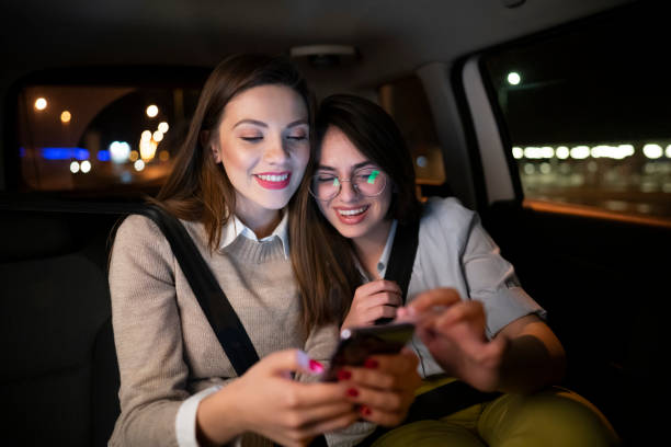 Two smiling women using a phone together while driving on the back seat of a car stock photo