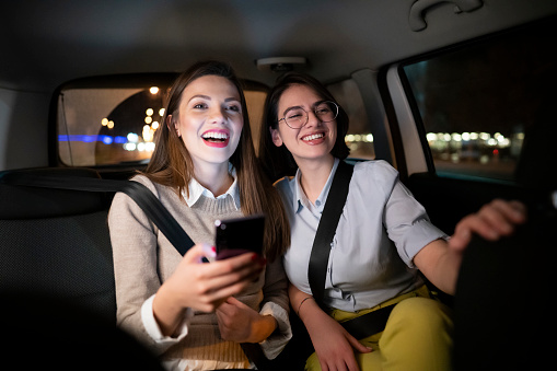 Two smiling Caucasian young women driving in the back seat of a car and using a smartphone together at night.