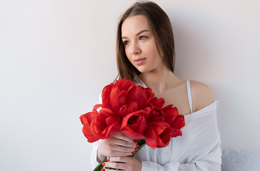Portrait of an attractive girl with a bouquet of red tulips in the morning in the bedroom for Happy Birthday. Caucasian woman with flowers on the bed close-up. White background.