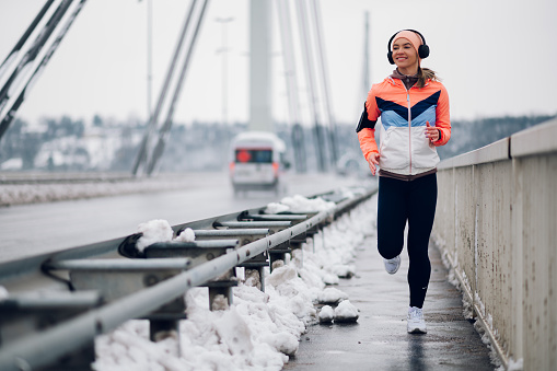 Beautiful young woman running and training on a bridge on a cold winter day.Winter fitness. Motivation concept. Urban city jogger. Outdoor fitness concept.