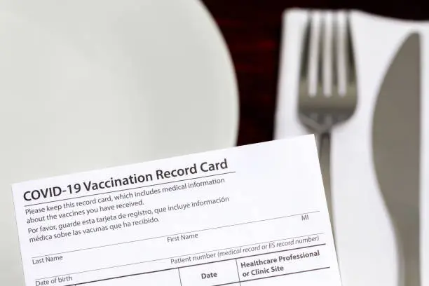 Photo of Covid-19 vaccination card and restaurant table. Indoor vaccine mandate, entertainment restrictions and vaccination requirement concept.