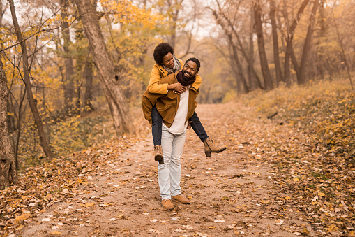 Happy African American man carrying his girlfriend in nature during autumn.