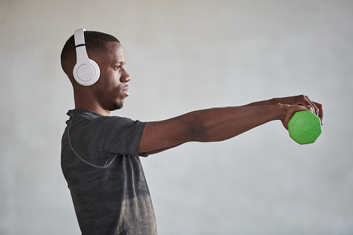 Horizontal medium side view shot of young Black man wearing white headphones listening to music and doing exercise with dumbbell