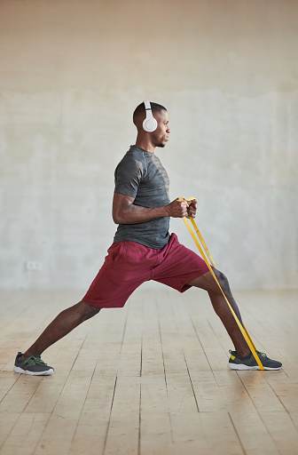 Vertical full shot of handsome young Black man doing strengthening exercise with resistance band and listening to music in headphones