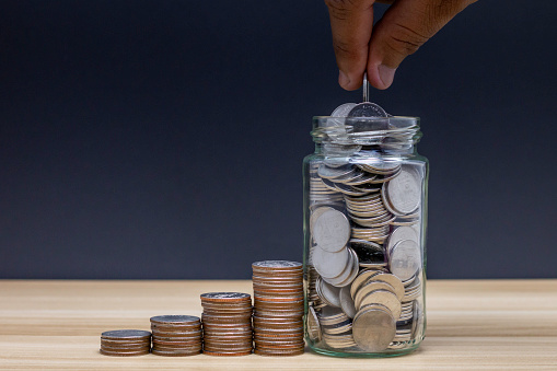 Saving money for sustainable life in the future concept: Coins in a clear glass jar with vintage clock nearby. Saving is money saved for education, paying taxes, insurance, health, travel.
