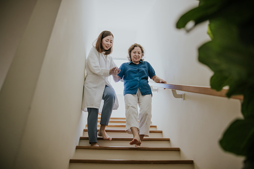 Asian grandmother and her young female medical physiotherapist while trying to moving down.