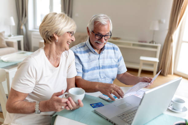 happy middle aged husband and wife sitting at table with laptop and paper bills, calculating domestic incomes together at home. - incomes imagens e fotografias de stock