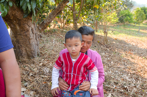 Candid portrait  of Thai farm women and a boy in shadow of trees in plantation area. The boy is wearing a traditional vest. Both are lookig at camera.  Scene is in countryside of Chiang Mai province