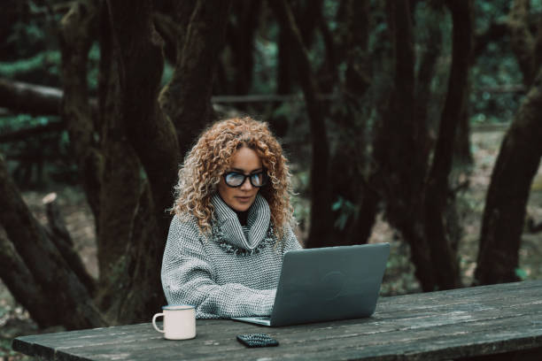 Curly long hair blonde adult woman use laptop computer outdoors sitting in the woods - concept of modern free female peoplelifestyle in smart and remote working activity - online business economy with phone and roaming stock photo