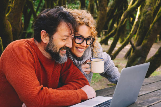 Happy modern couple use together laptop computer outdoors with trees and woods in background - video call and smart remote working activity lifestyle concept with cheerful people and technology